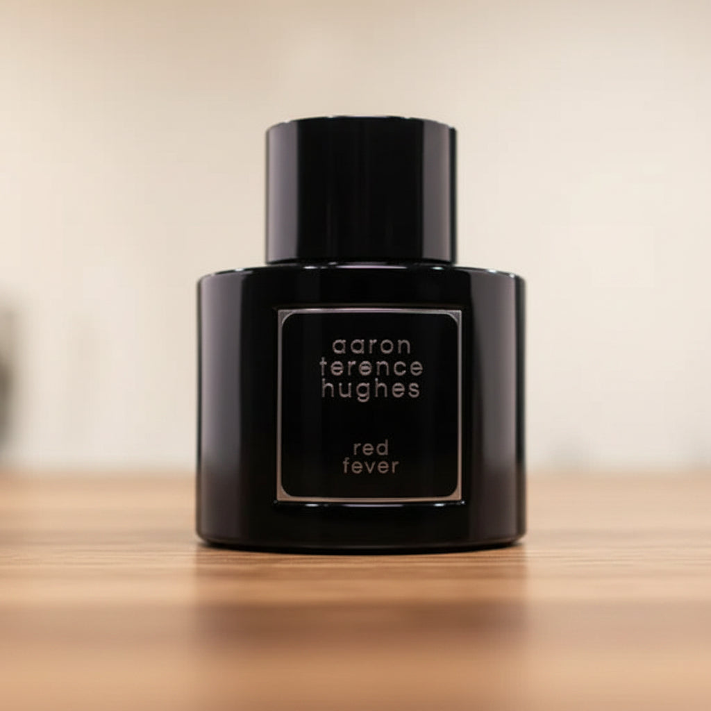 Black perfume bottle labeled 'aaron terence hughes red fever' on a reflective surface with a blurred background.