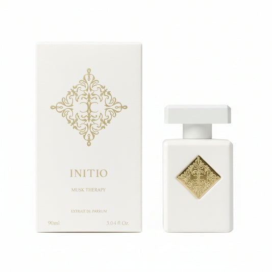 A white rectangular perfume bottle with a gold diamond emblem stands next to its matching white box, which features a gold ornate pattern and the text 'INITIO MUSC THERAPY EXTRAIT DE PARFUM'.